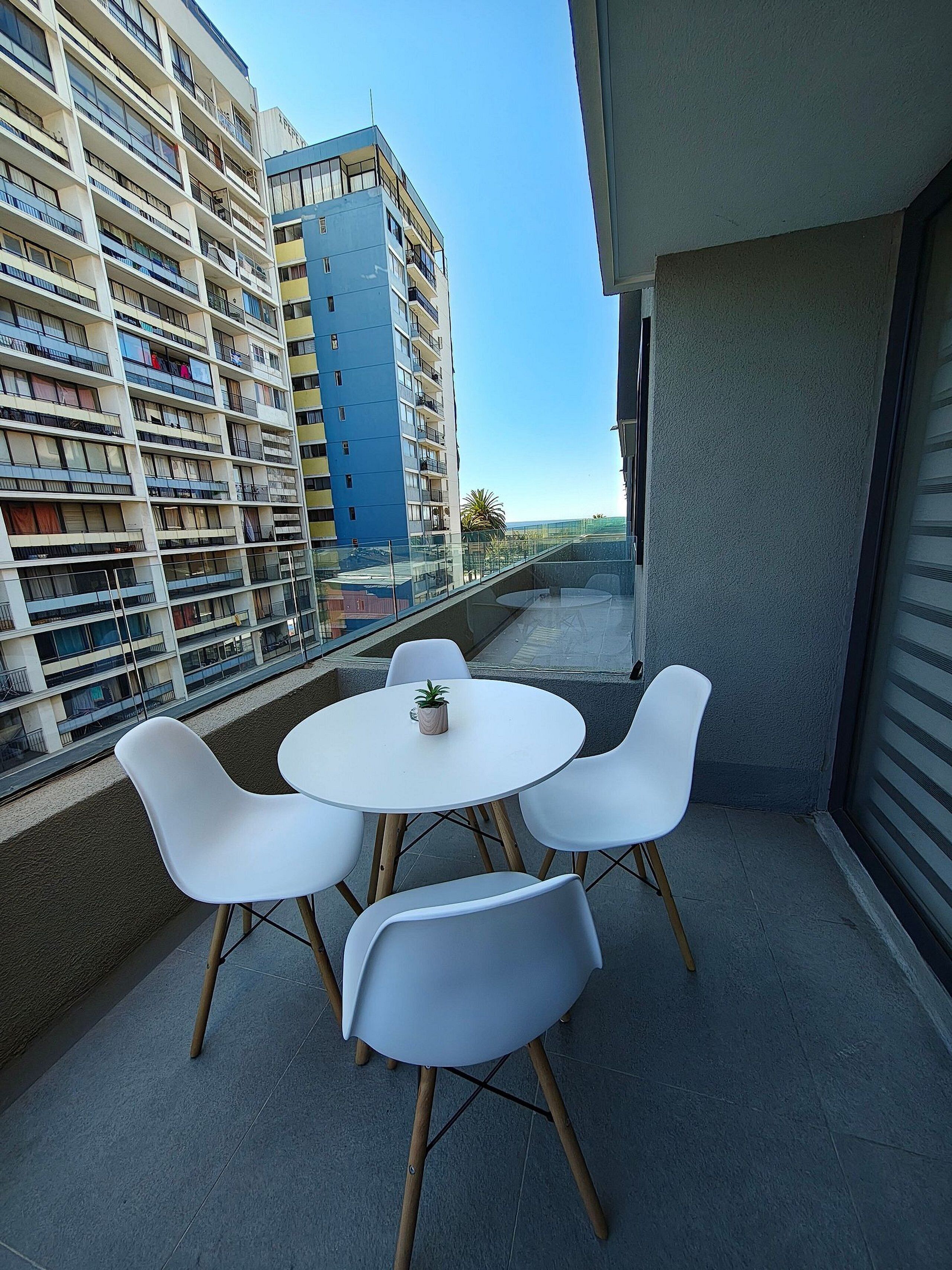 Un balcón con una mesa redonda y cuatro sillas blancas, con vistas a un paisaje urbano de edificios altos.