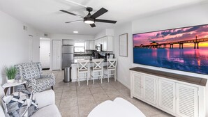 Un salon moderne doté d’un ventilateur de plafond, d’un grand écran de télévision diffusant une vue sur un coucher de soleil, d’un coin cuisine avec des meubles blancs et d’un coin repas avec des chaises blanches.