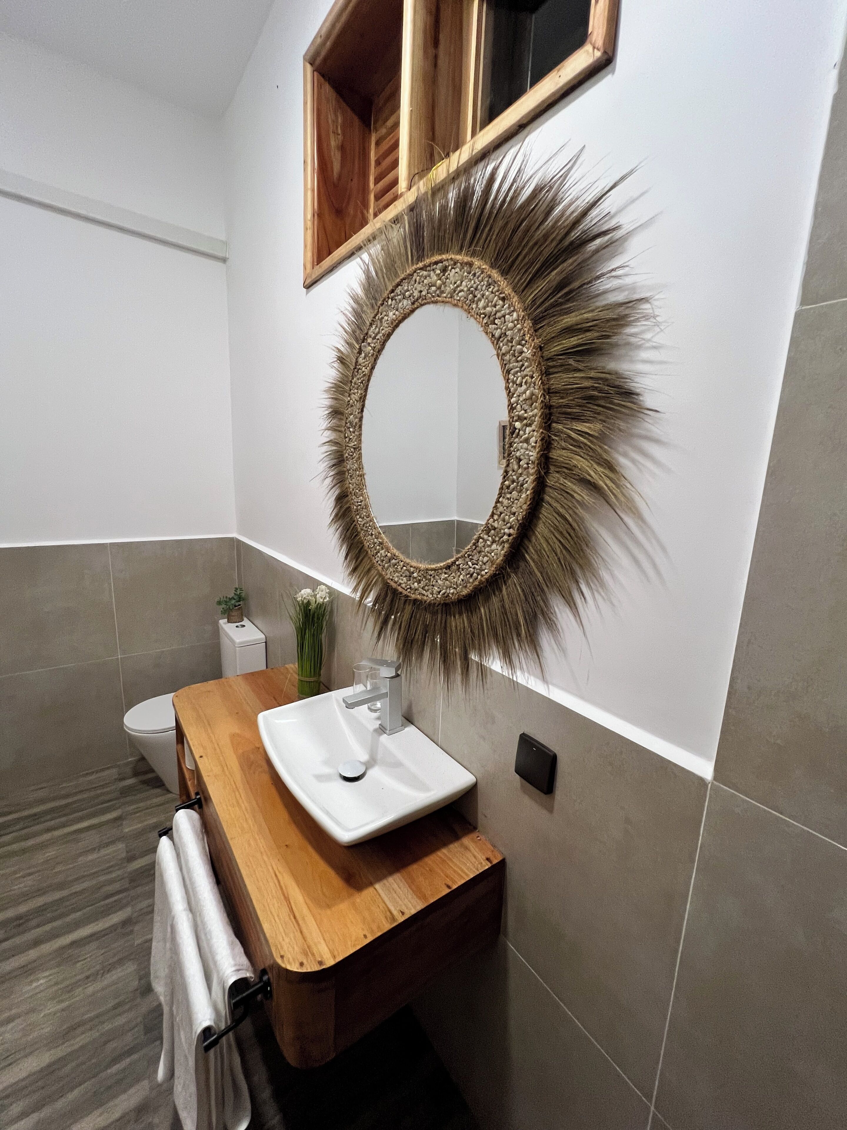 Une salle de bain avec un meuble-lavabo en bois, un lavabo blanc, un miroir avec un cadre décoratif, des toilettes et une petite plante.