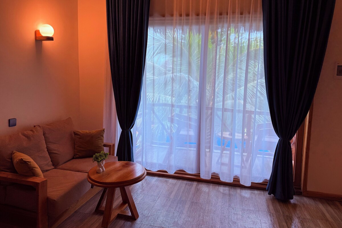 A cozy living room with a sofa, a small wooden table, and a large window with sheer curtains.