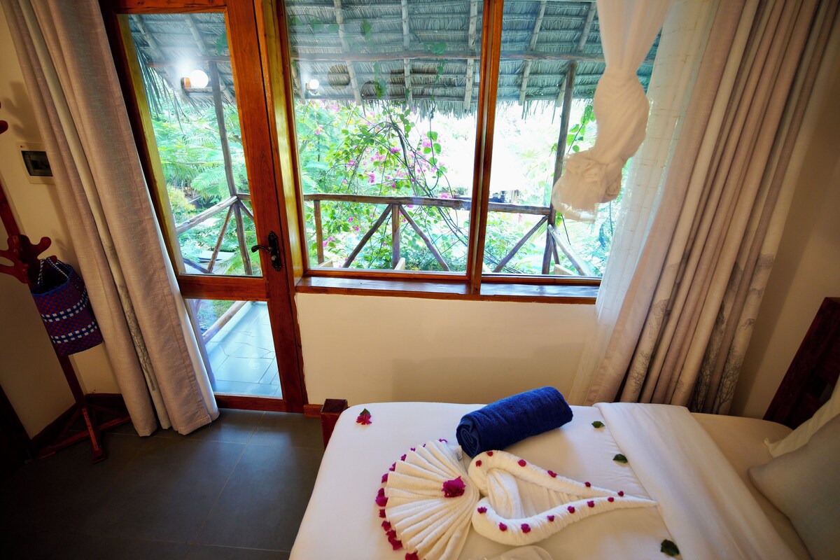 A hotel room with a bed, a window with curtains, and a view of greenery outside.