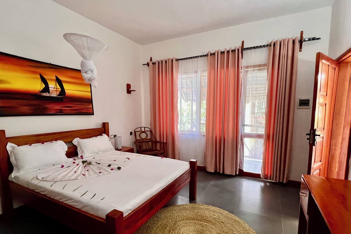 Un dormitorio con cama, una silla, un cuadro, cortinas y una puerta.