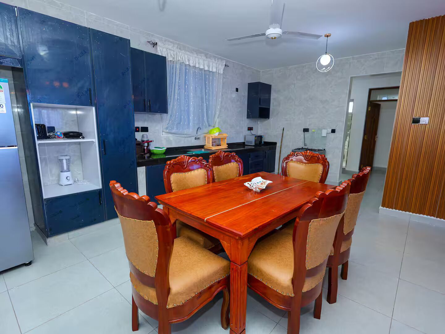 A dining area with a wooden table and six chairs, a kitchen with a refrigerator, a blender, and a microwave, and a ceiling fan.
