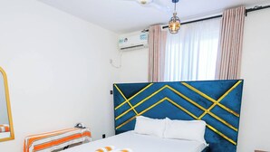 A bedroom with a bed, a ceiling fan, an air conditioning unit, a window with curtains, and a small wooden table. - Homes by Pesh - Nyali, 2 bedroom - Mombasa. (el doret)