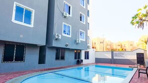 A swimming pool with a tiled deck, surrounded by a building with multiple windows and air conditioning units. - Homes by Pesh - Nyali, 2 bedroom - Mombasa. (el doret)