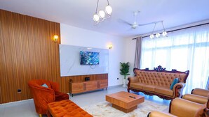 A modern living room with a wooden accent wall, a large flat-screen TV, a brown sofa, and orange armchairs. - Homes by Pesh - Nyali, 2 bedroom - Mombasa. (el doret)