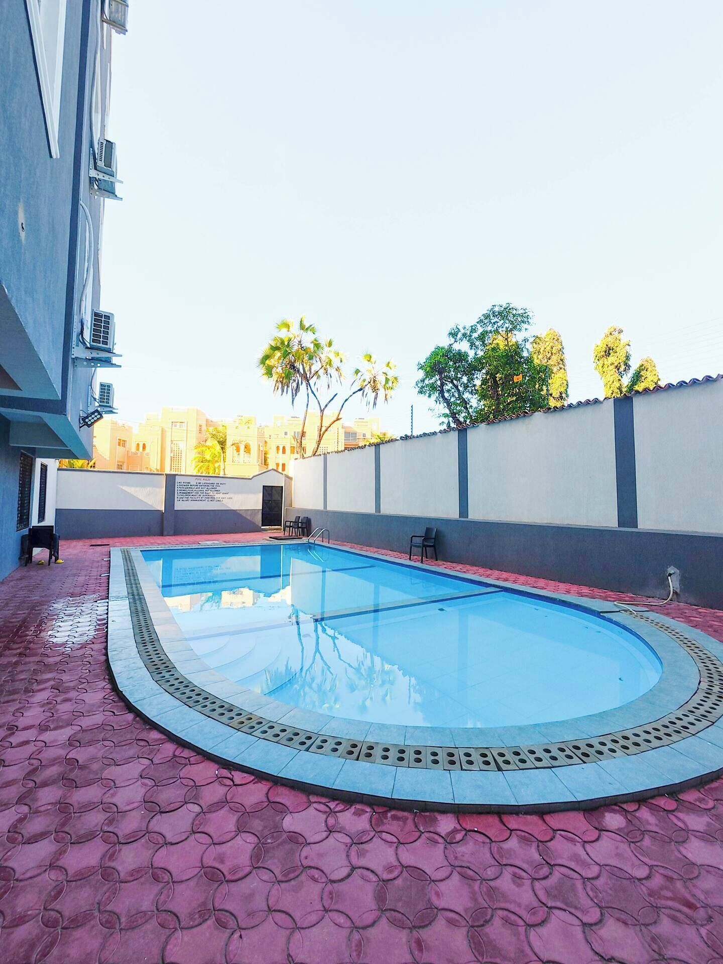 A swimming pool with a red patterned floor, surrounded by a low wall and a building.