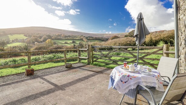 A patio with a table set for two, overlooking a scenic countryside view. - Swallow Cottage (South Brent)