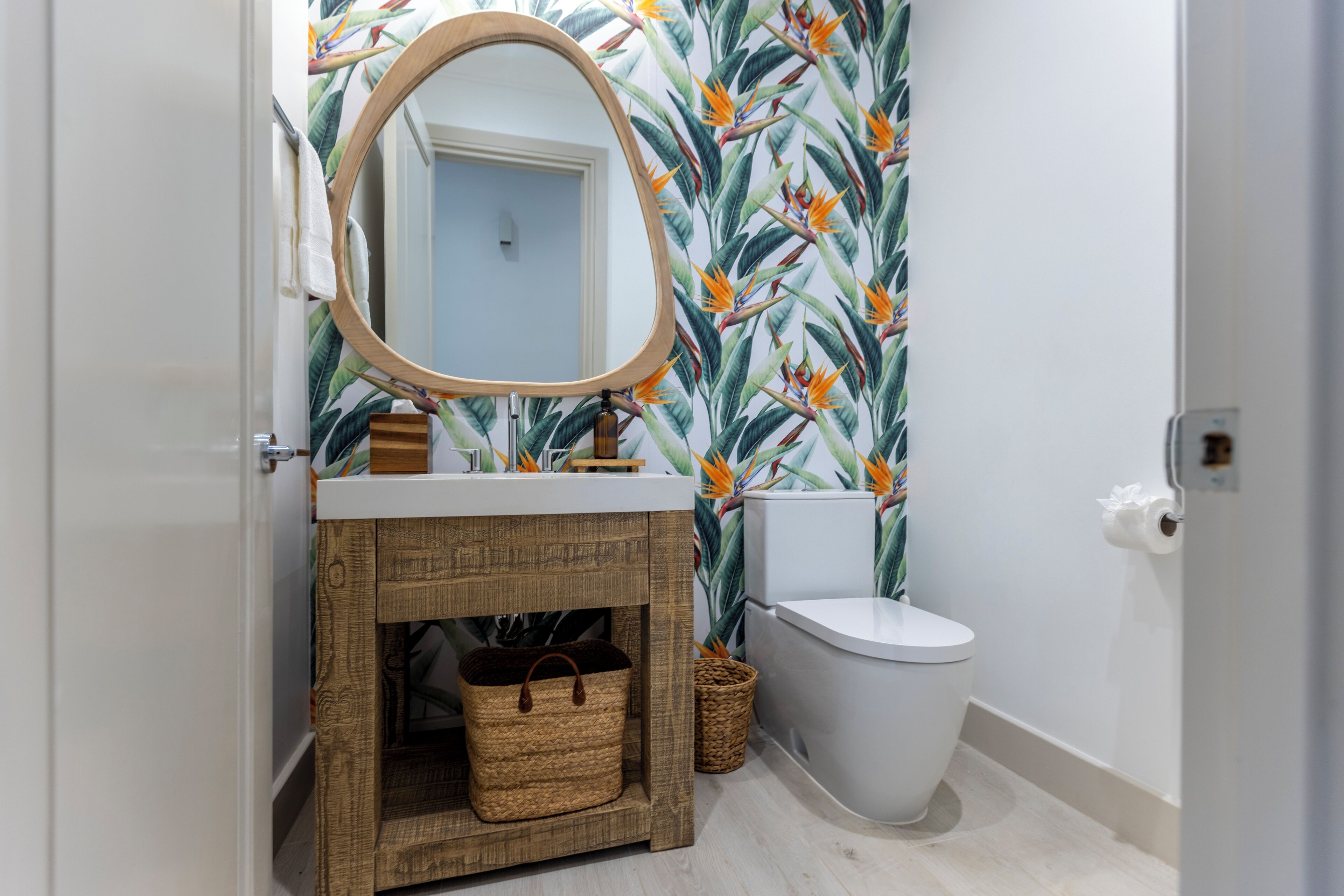 A bathroom with a wooden vanity, a round mirror, a toilet, and a wall with a tropical leaf pattern.