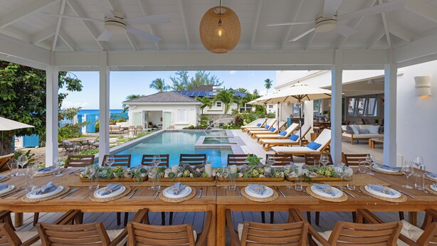 A covered outdoor dining area with a pool, lounge chairs, and a view of a house and palm trees. - Kaiso Beachfront Villa (Prospect)