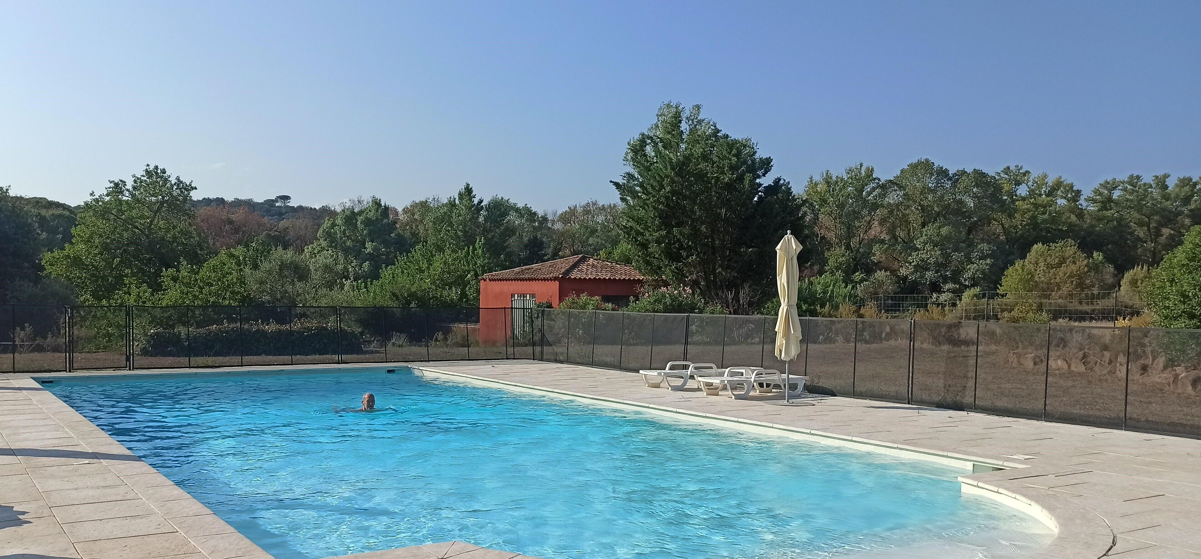 Ein Schwimmbecken mit gefliestem Beckenrand, ein rotes Gebäude im Hintergrund und ein Zaun mit Bäumen und Hügeln in der Ferne.