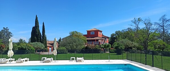 En swimmingpool med liggestole og parasoller, omgivet af hegn og grønt, med en rød bygning i baggrunden.