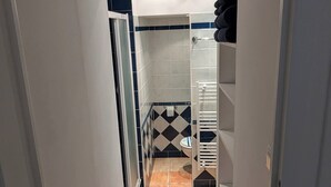Um corredor estreito com piso ladrilhado que leva a um banheiro com vaso sanitário e chuveiro.