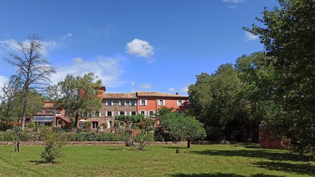 Um edifício grande e vermelho, com várias janelas e varandas, cercado por vegetação exuberante e um céu azul límpido.