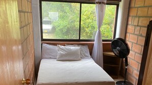 A bedroom with a bed, a window with a view of greenery, a standing fan, and a brick wall. - FARM WITH BEAUTIFUL VIEW 40MIN FROM MEDELLÍN. SPORTS COURT, SWIMMING POOL AND GYM. (Barbosa)