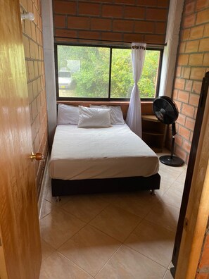 A bedroom with a bed, a window with a view of greenery, a standing fan, and a brick wall. - FARM WITH BEAUTIFUL VIEW 40MIN FROM MEDELLÍN. SPORTS COURT, SWIMMING POOL AND GYM. (Barbosa)