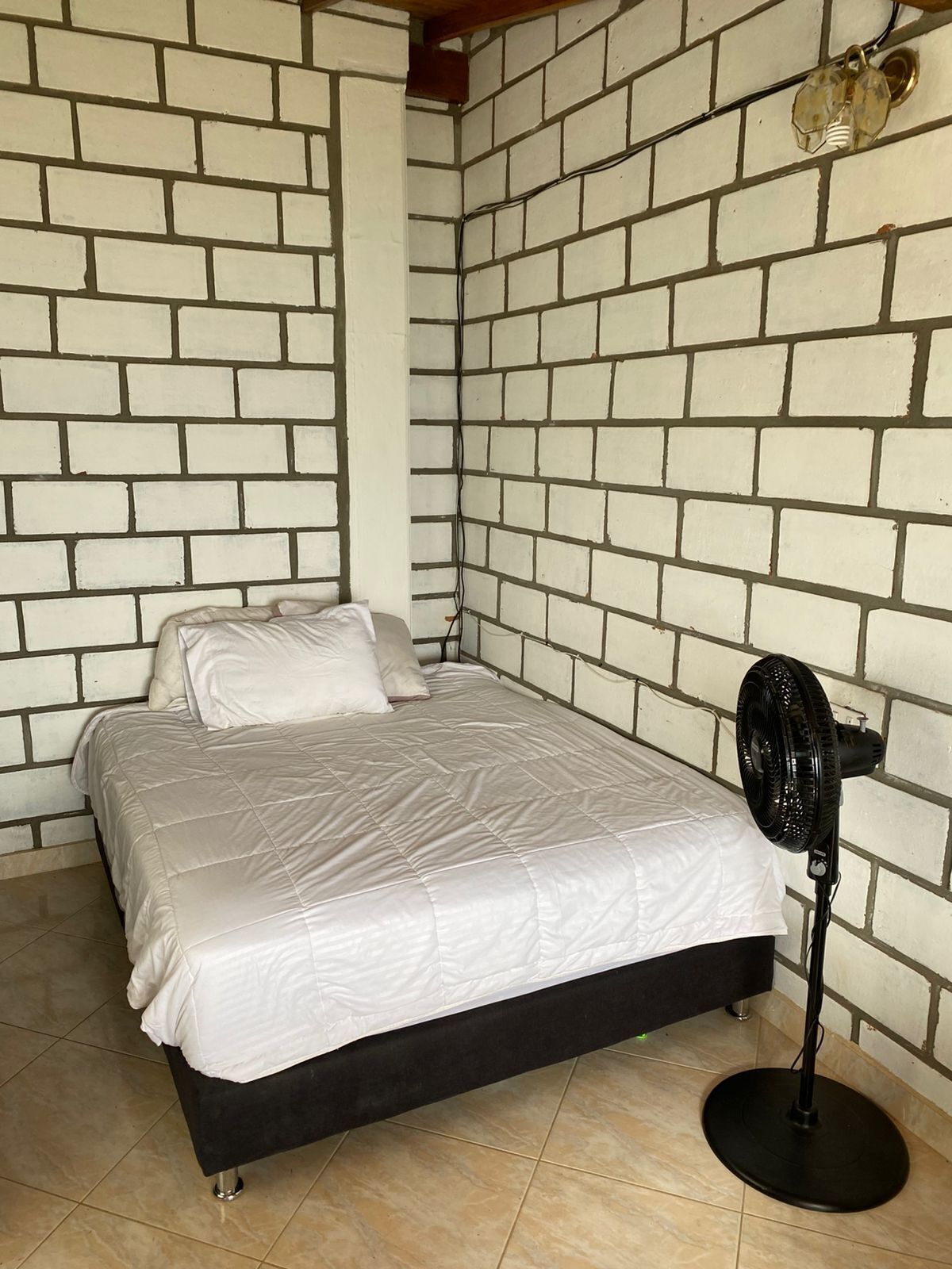 Una habitación con una cama frente a una pared de ladrillo, un ventilador de pie y piso de baldosas.