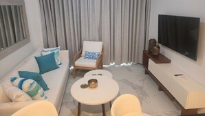 Una sala de estar moderna con un sofá, sillones, una pequeña mesa redonda, un televisor y un amplio ventanal con cortinas.