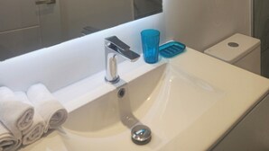 Un baño con lavamanos blanco, un espejo, toallero con toallas, un vaso de vidrio azul y un inodoro.