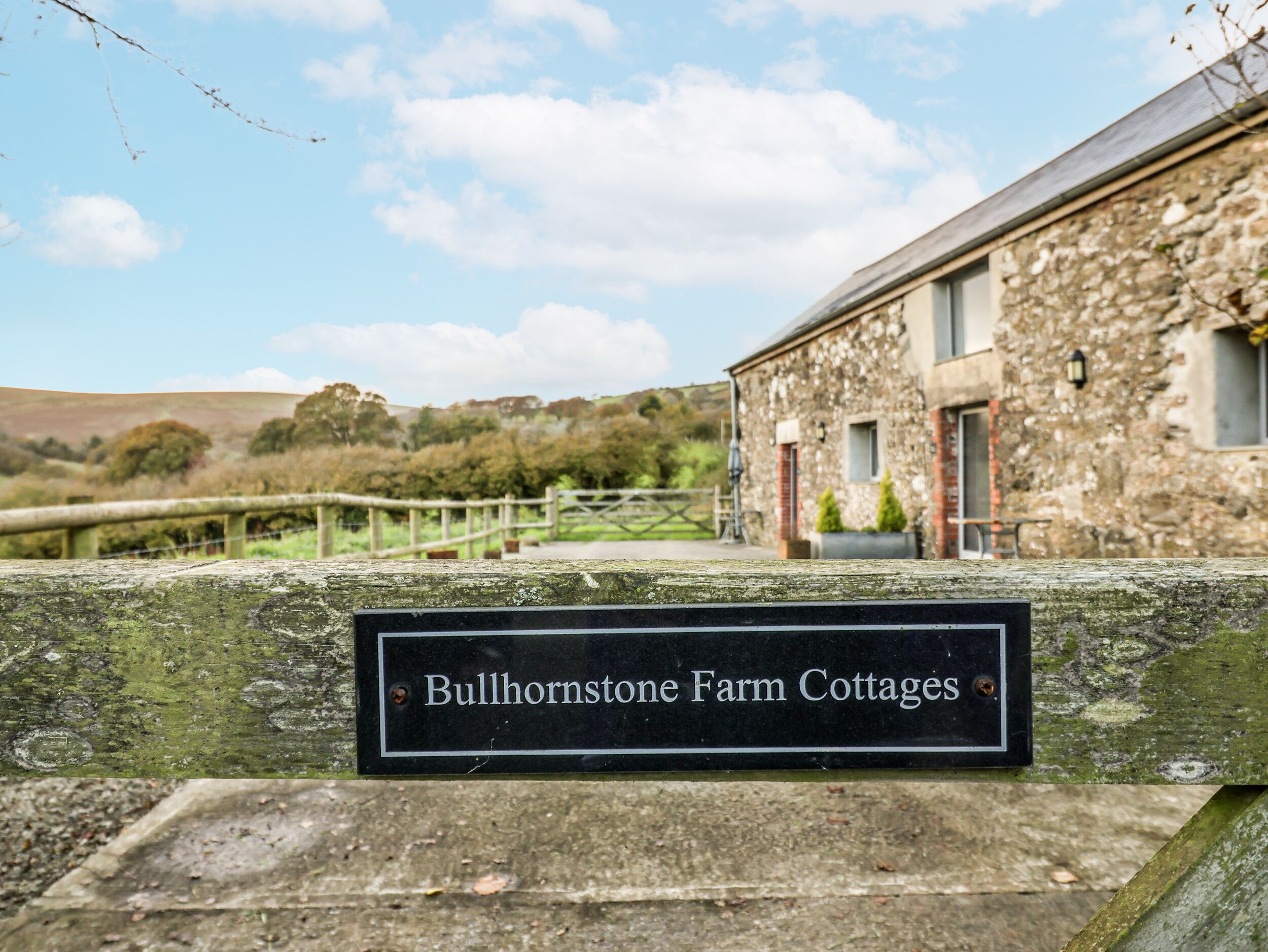 A rustic stone building labeled "Bullhornstone Farm Cottages" with a wooden fence and a clear sky in the background.