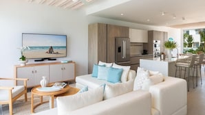 A modern living room with a sofa, coffee table, and TV. A kitchen with a fridge, oven, and bar area is visible. - Turks & Caicos Luxury | Oceanfront Boathouse Villa w/ Private Dock & Sea Views (Long Bay Hills)