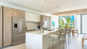 A modern kitchen with a large island, stainless steel appliances, and a dining area with wicker chairs. - Turks & Caicos Luxury | Oceanfront Boathouse Villa w/ Private Dock & Sea Views (Long Bay Hills)