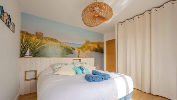 A bedroom with a large bed, a wall-mounted painting of a beach scene, a woven light fixture, and a wooden headboard. - The Villa of the Waves (Châtelaillon-Plage)