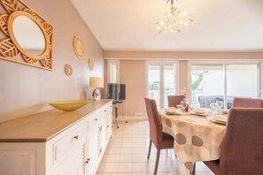 A dining area with a table set for four, a television on a stand, and a large window with a view of greenery outside. - Apartment - The Gentle Tides (La Rochelle)