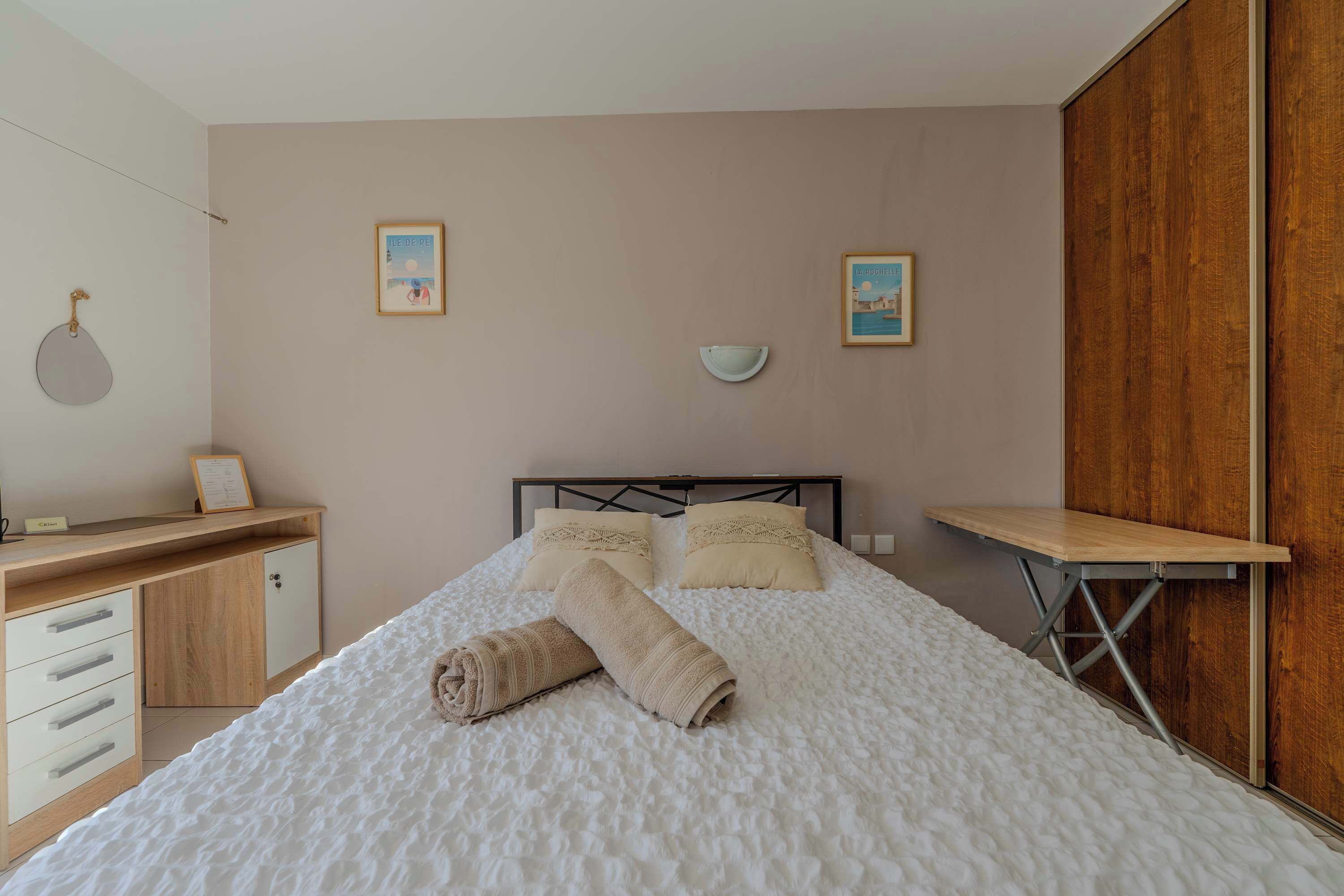 A neatly made bed with a rolled towel, a wooden desk, a wall-mounted bowl, and two framed pictures on the wall.