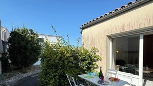 An outdoor patio with a table set for dining, surrounded by greenery and a pathway leading to a building with a tiled roof. - Golden Waves Aparthotel - Kiwi Residence (Aytré)