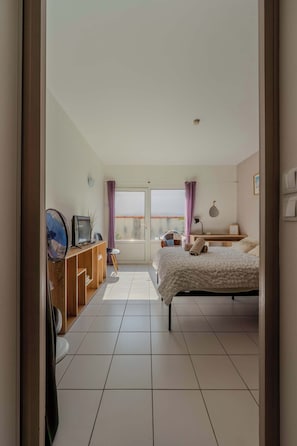 Ein Schlafzimmer mit einem Bett, einem Schreibtisch, einem Stuhl, einem Ventilator und einem Fenster mit Vorhängen.