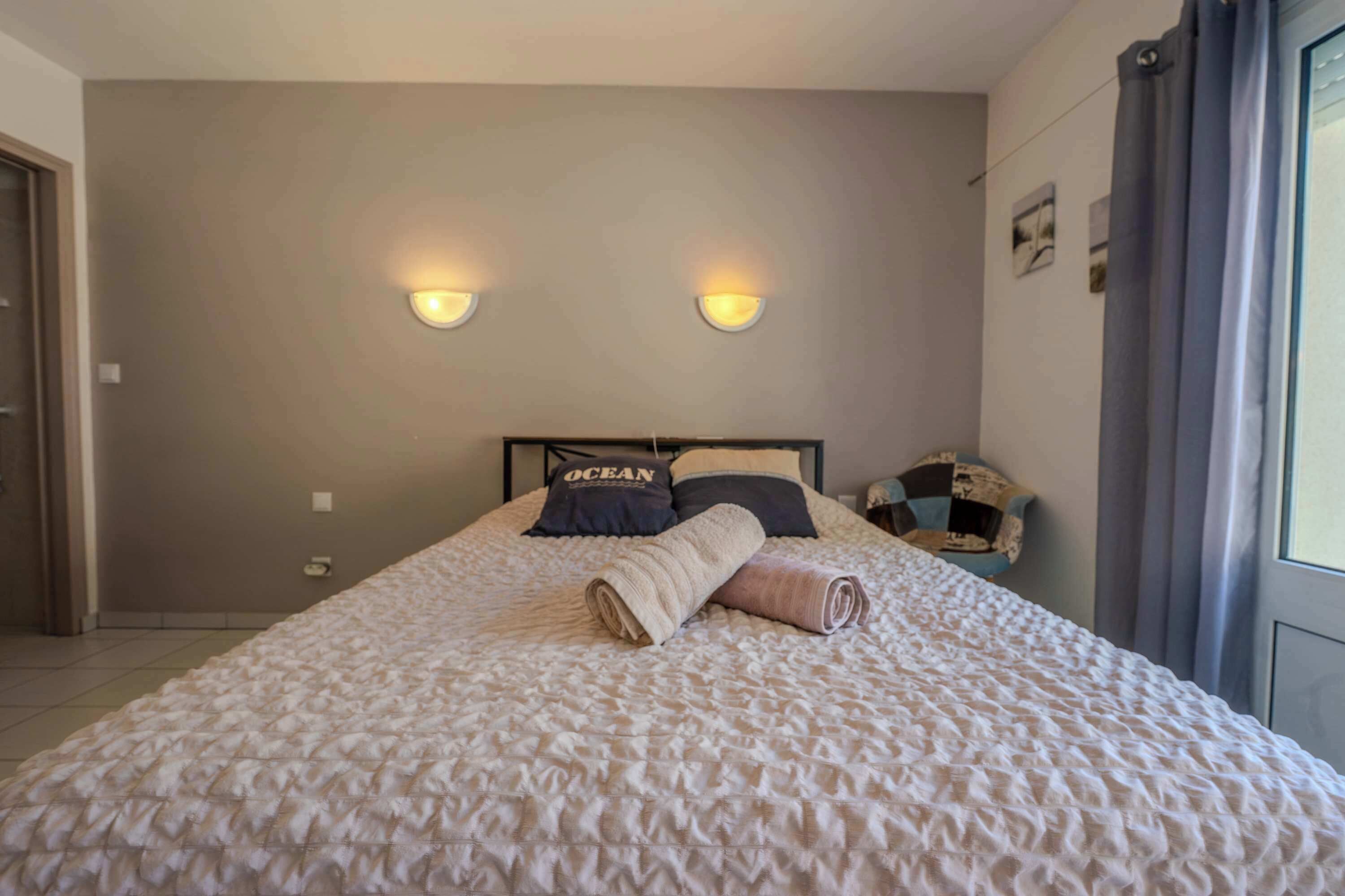 Un lit bien fait, recouvert d’une couverture à motifs, avec deux lampes fixées au mur et une tête de lit ornée d’un coussin sur lequel est inscrit le mot « OCÉAN ».
