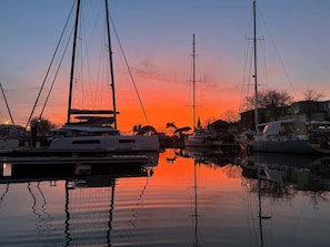 En småbåtshamn med segelbåtar förtöjda, stilla vatten som speglar båtarna och en solnedgång i bakgrunden.