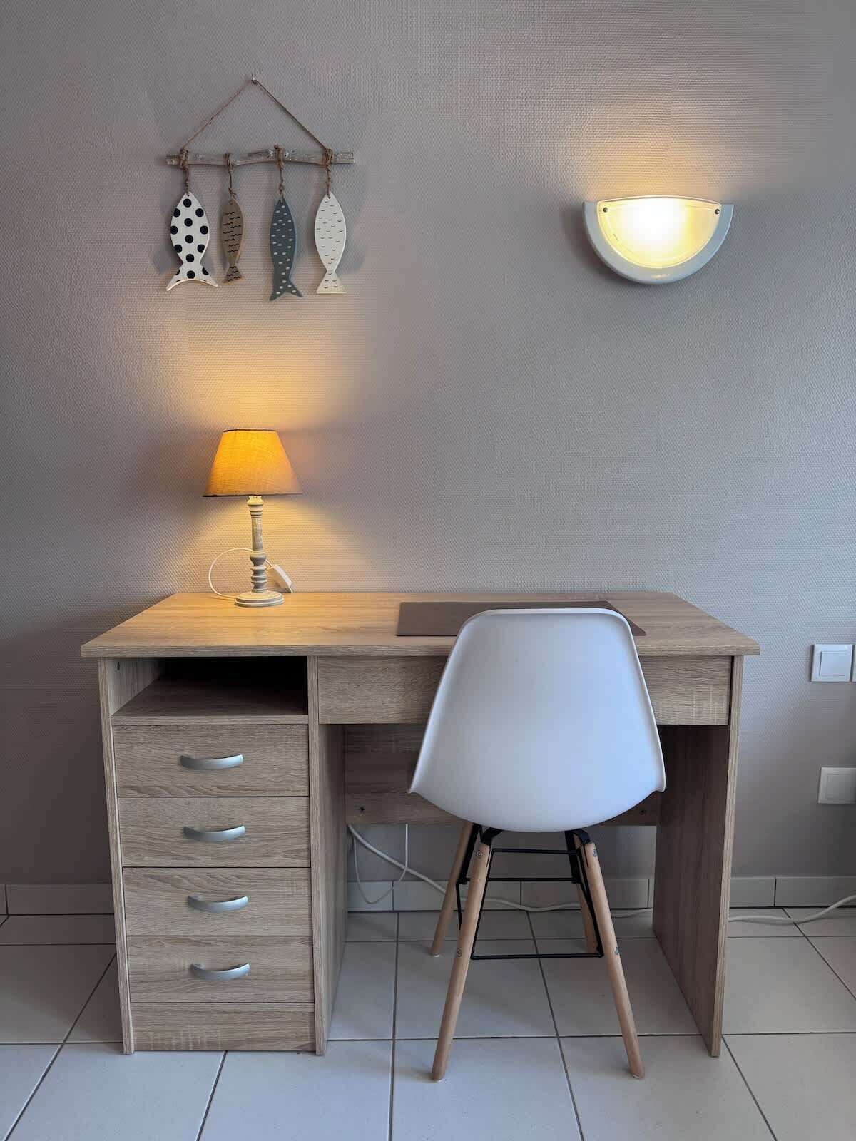 Ett skrivbord i trä med en vit stol, en lampa och dekorationer som hänger på väggen.