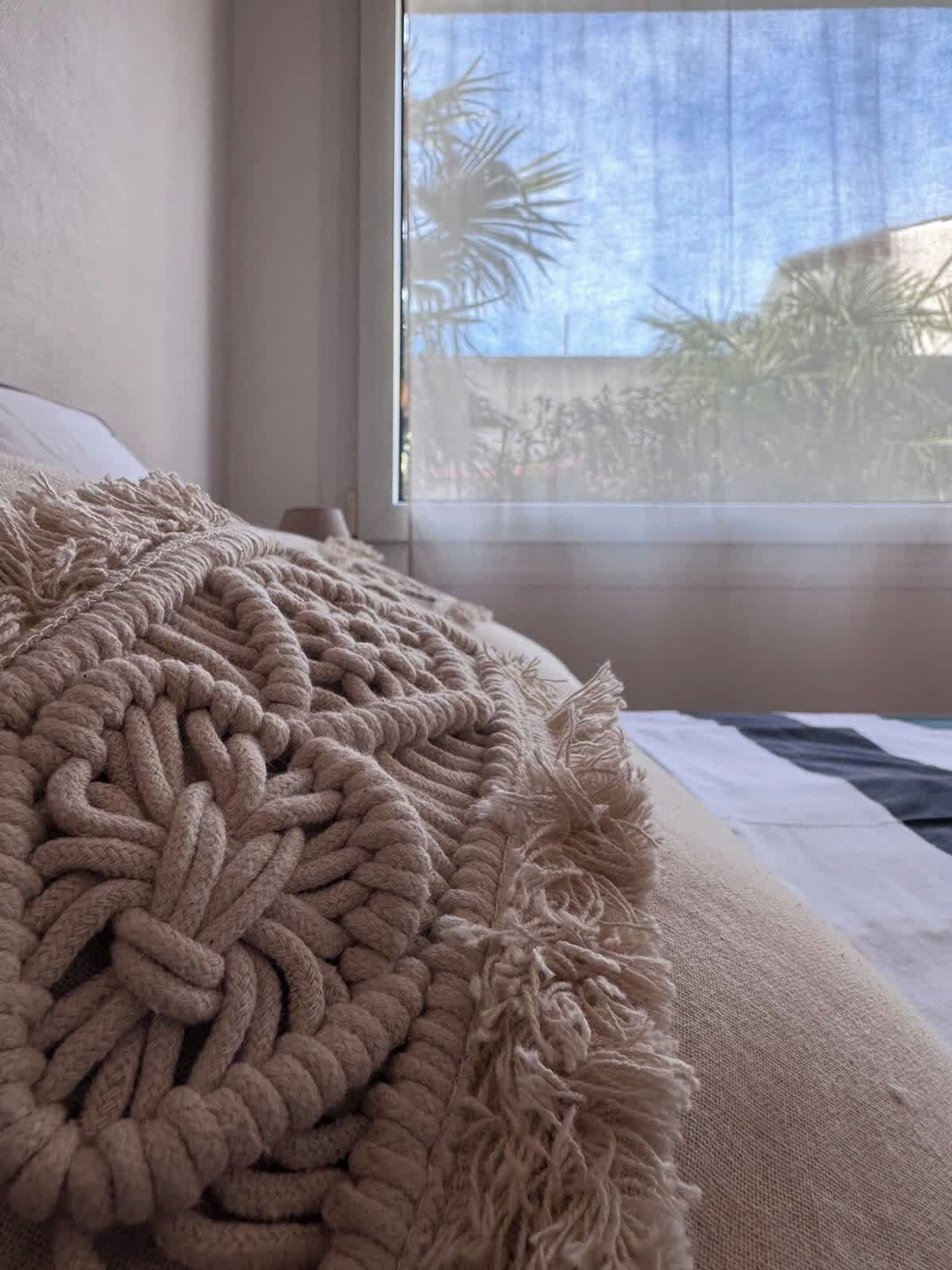 A bed with a textured beige blanket and a view of palm trees outside the window.