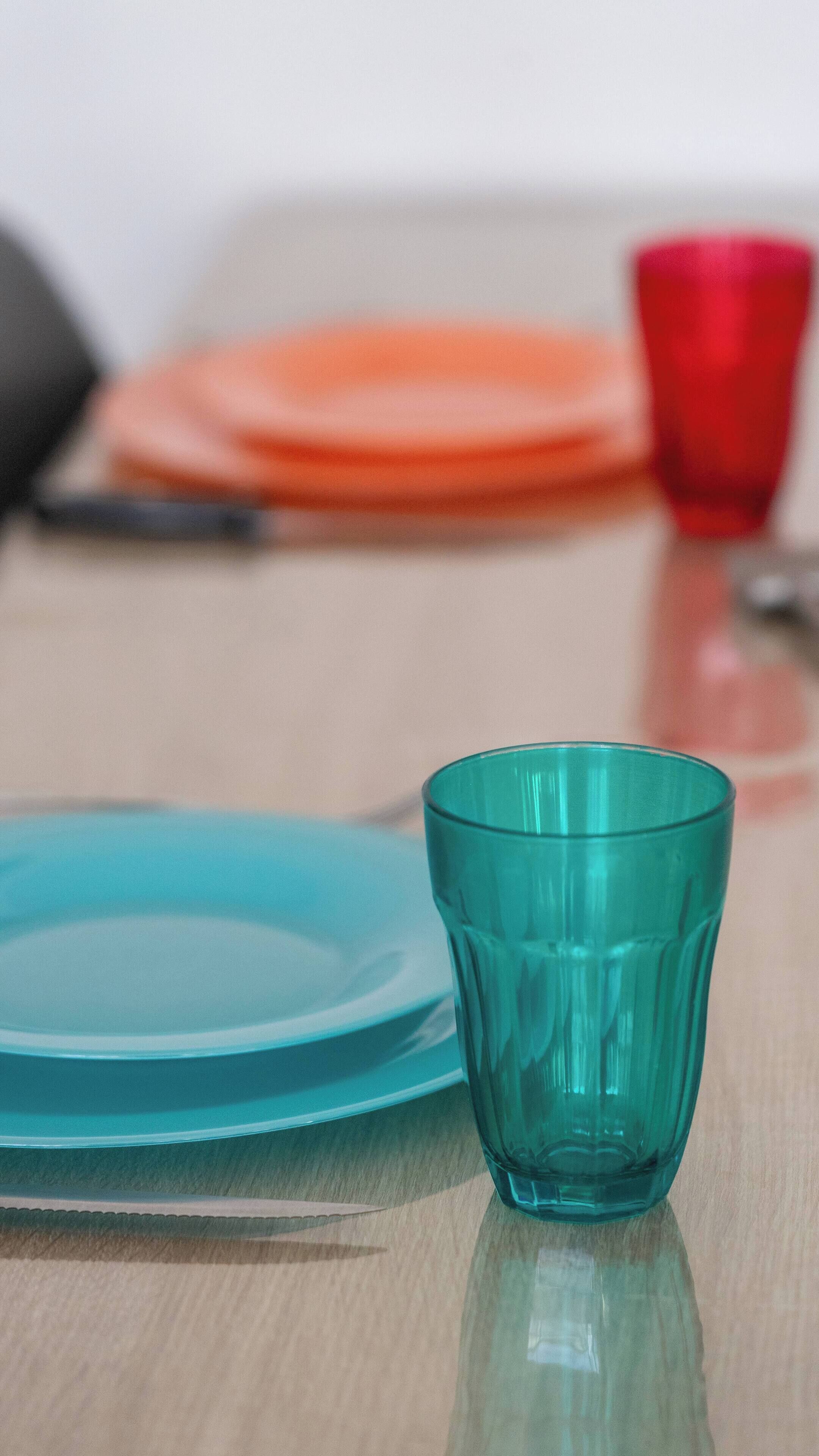 Et turkis glas på et bord med en blå tallerken og en rød kop i baggrunden.