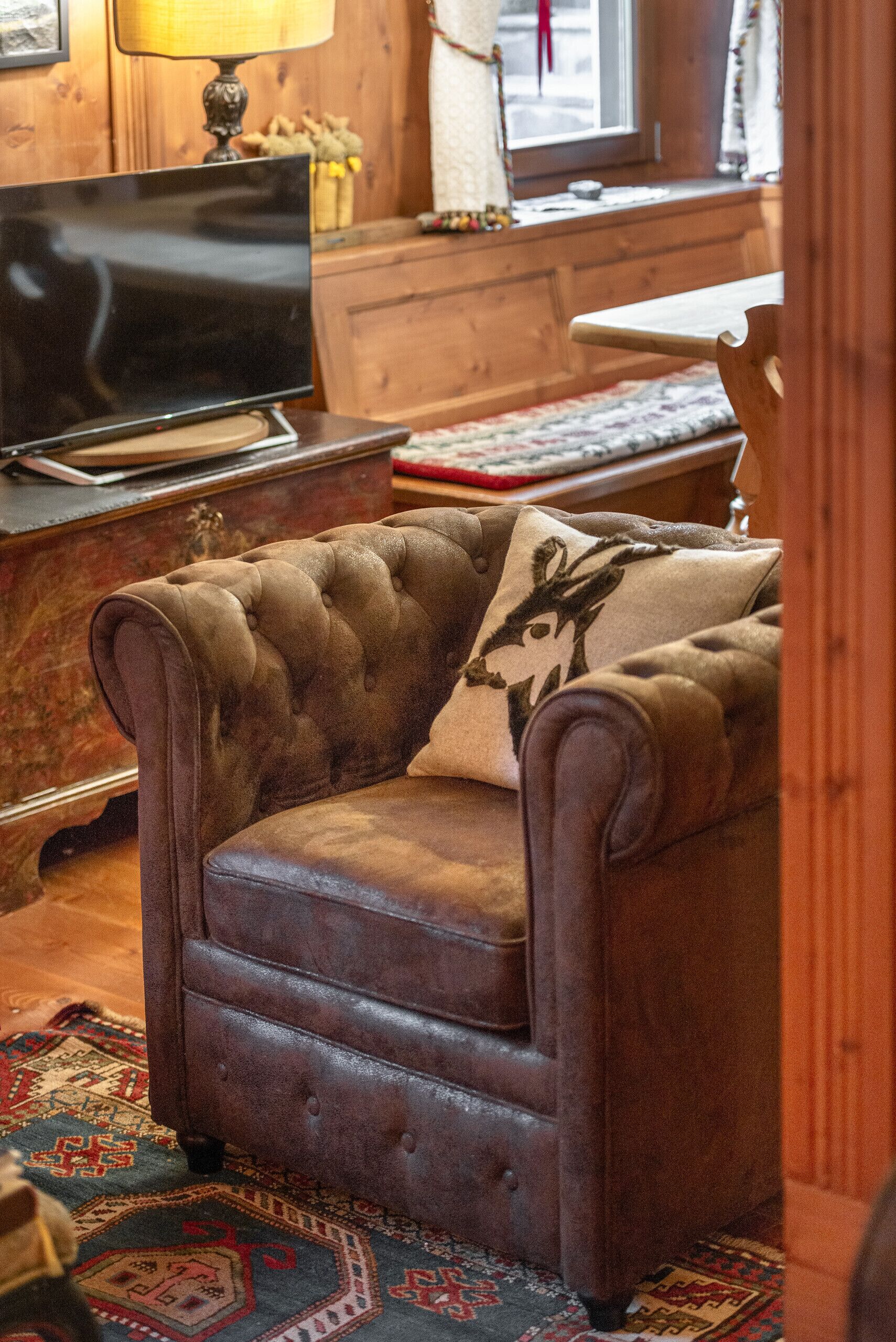 Una poltrona in pelle marrone con un cuscino decorato, un mobiletto in legno, un televisore e una finestra con tende.