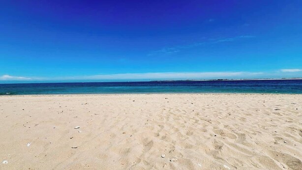 En bred, sandet strand med klart, blåt vand og en fjern horisont.