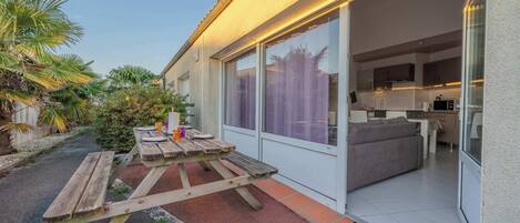 Een modern huis met een patio, een keuken zichtbaar door glazen deuren, en een kleine tuin met palmbomen.