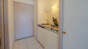 Un couloir étroit avec une porte fermée, un coin cuisine avec un évier et une cuisinière, et un sol carrelé.