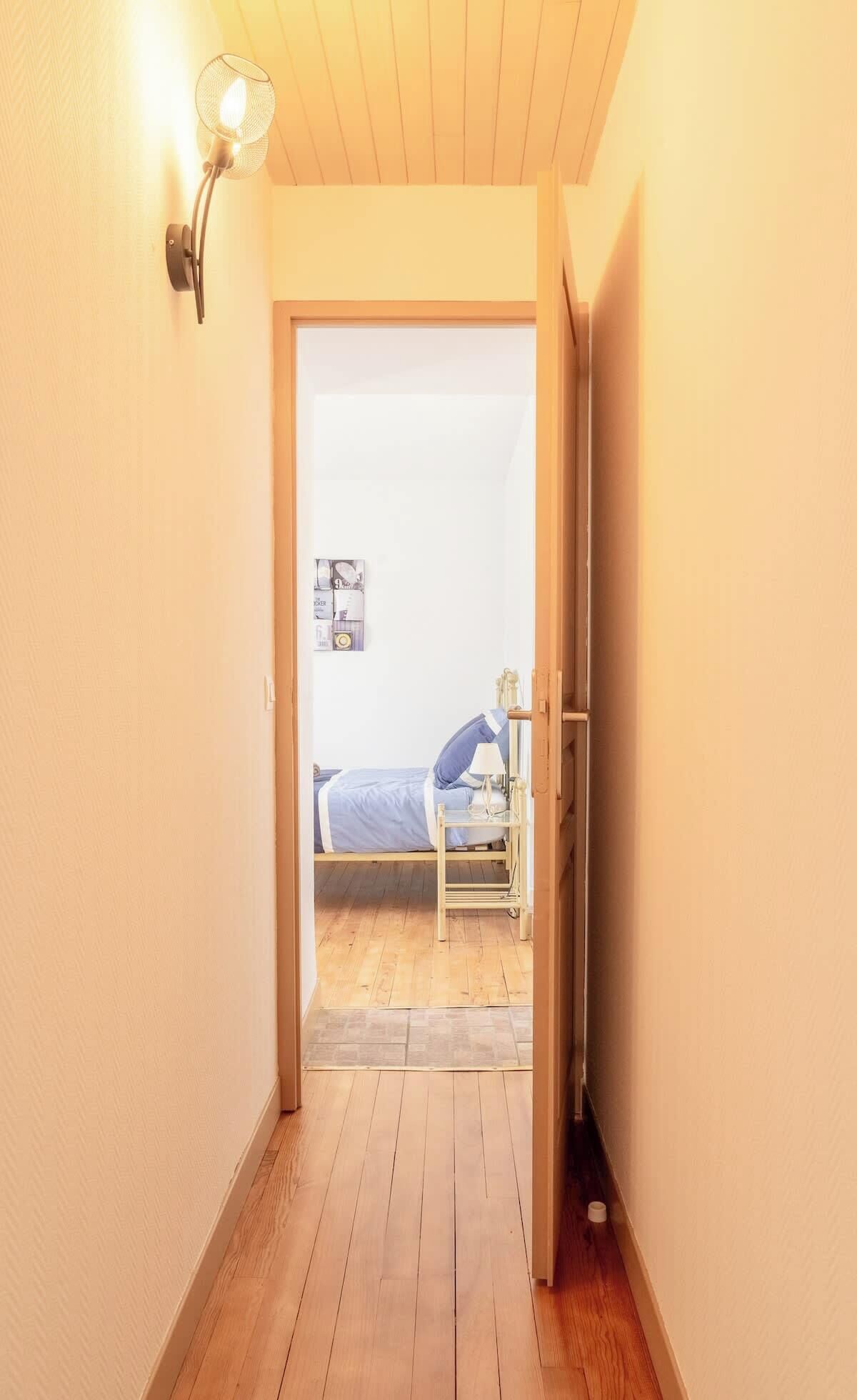 En smal gang der fører til et soveværelse med en seng, et natbord og en lampe på væggen.