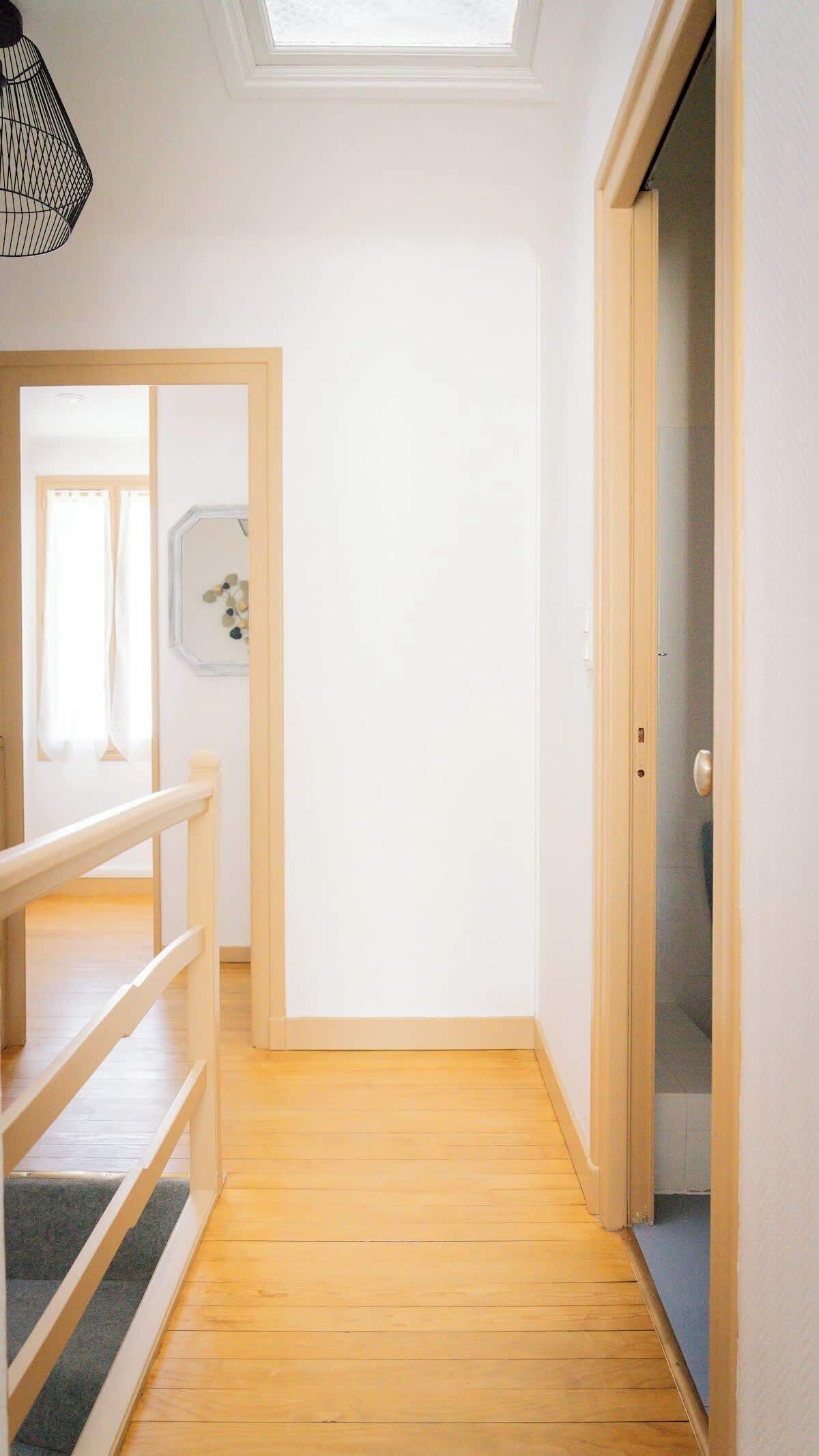 Un couloir étroit avec un plancher en bois, une lucarne et une porte donnant sur une pièce.