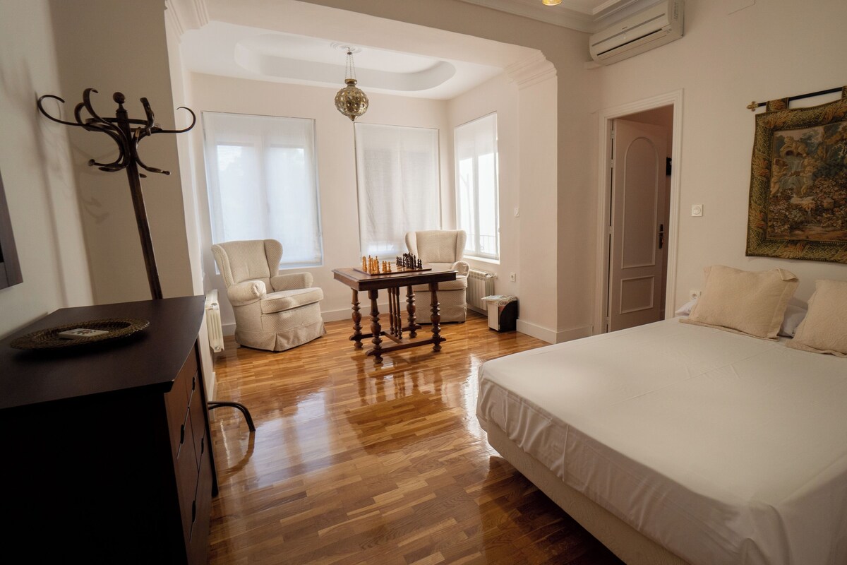 A bedroom with a large bed, a wooden table with chess pieces, two chairs, a coat rack, and a painting on the wall.