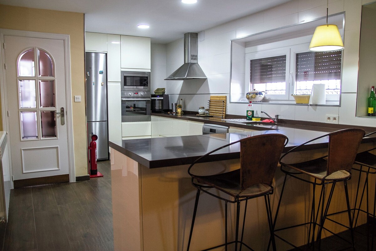 A modern kitchen with a central island, bar stools, a stainless steel refrigerator, and a large window with blinds.