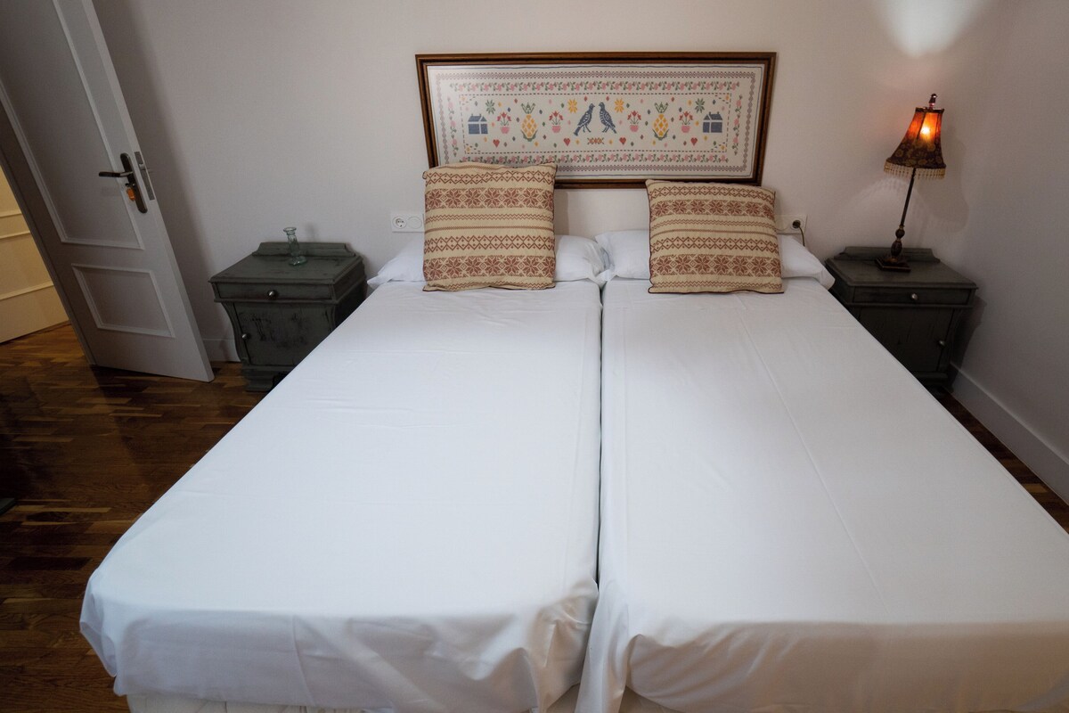 A double bed with white linens, two patterned pillows, a framed tapestry above the bed, and a bedside lamp on the right.