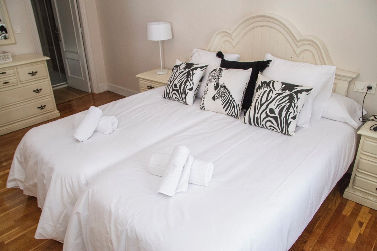 A neatly made bed with white bedding and zebra-patterned pillows, a bedside table with a lamp, and a wooden floor.