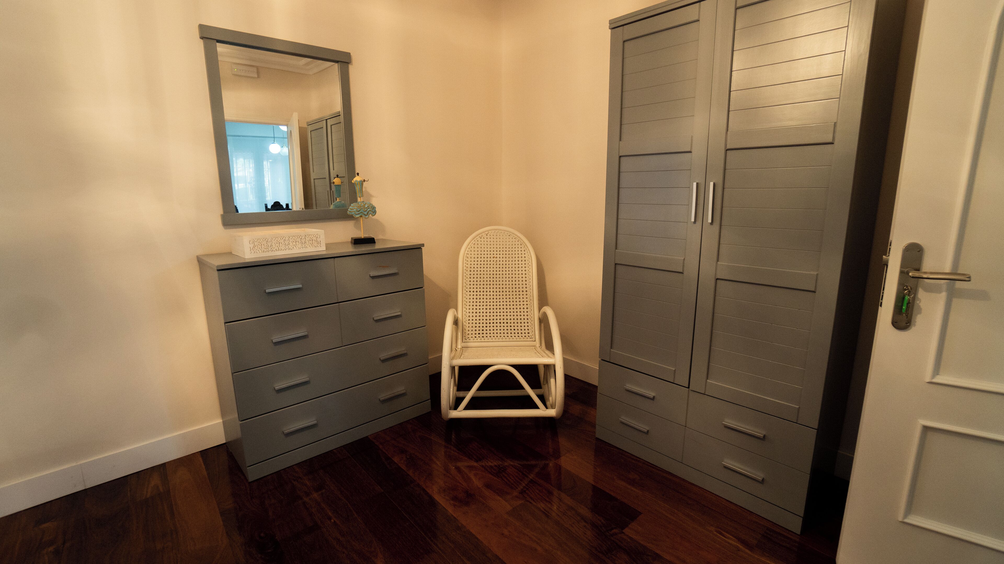 A small, narrow room with a wooden floor, a white chair, a dresser with drawers, a mirror, and a wardrobe.