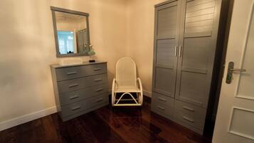 A small, narrow room with a wooden floor, a white chair, a dresser with drawers, a mirror, and a wardrobe.