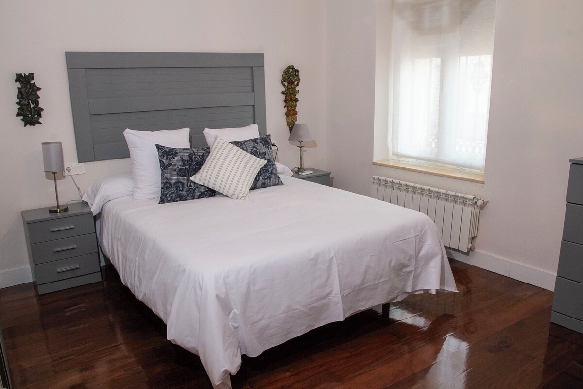 A bedroom with a large bed, grey headboard, white bedding, two bedside tables with lamps, a radiator, and a window with curtains.