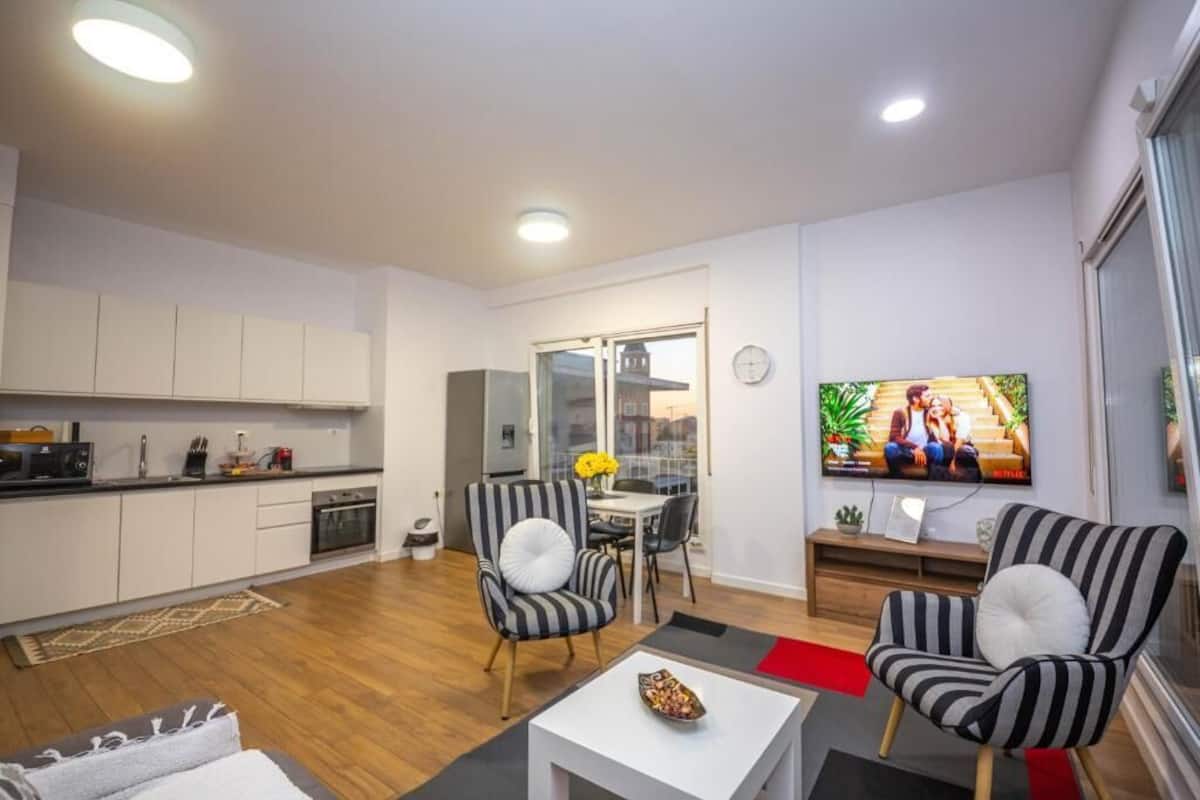 Un apartamento moderno con cocina, comedor y sala de estar. Hay un televisor montado en la pared, una mesa de comedor con sillas y dos sillones a rayas.